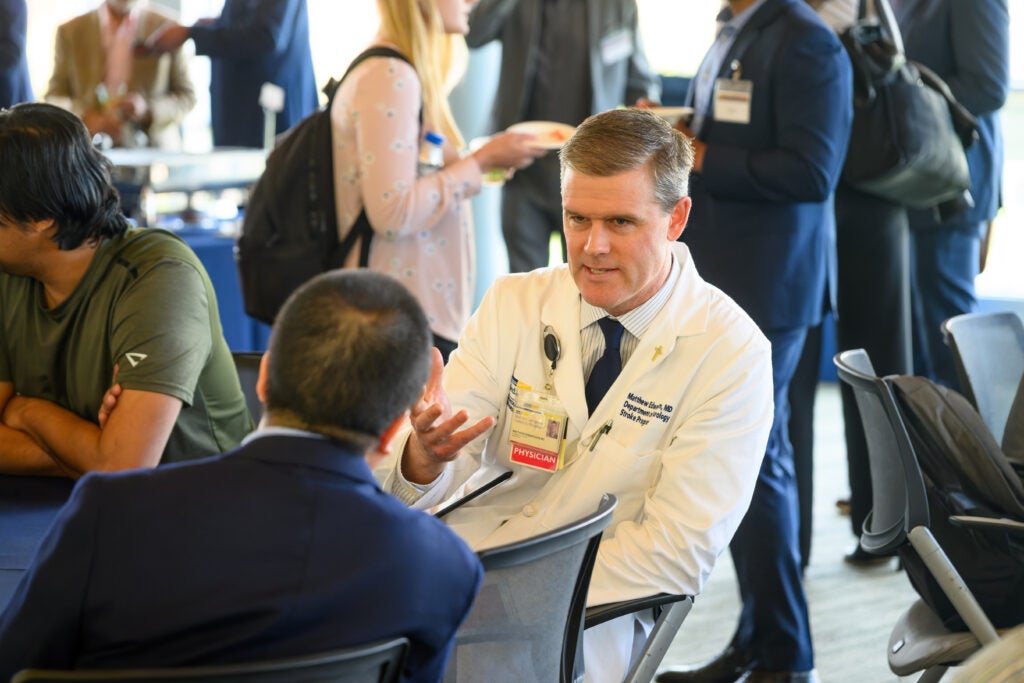 People sitting at a table, one in white coat, conversing
